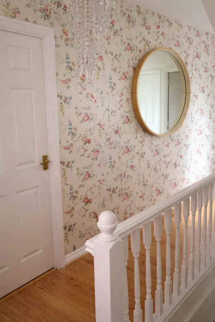 ممر داخلي مزين بورق جدران بنقوش زهرية ناعمة بطابع ريفي، يتناسق مع الأبواب البيضاء والدرابزين الخشبي، مع مرآة دائرية