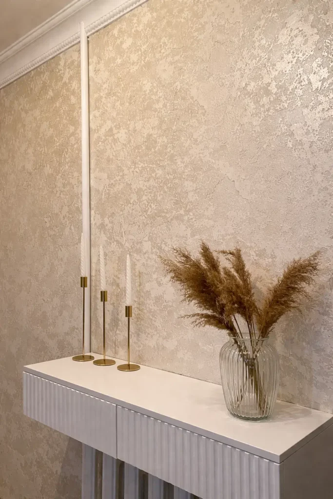 جدار مغطى بورق حائط بلون بيج زخرفي، أمامه طاولة بيضاء تعلوها شموع بحوامل ذهبية ومزهرية زجاجية تحتوي على نباتات جافة