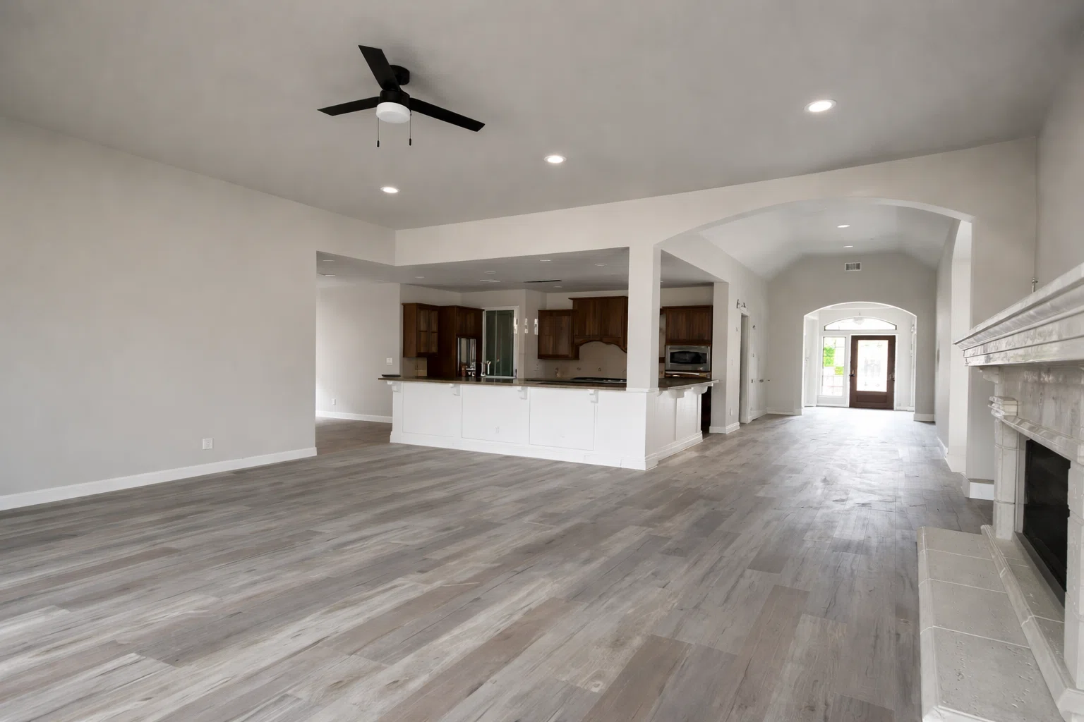 غرفة معيشة واسعة مع مطبخ أمريكي، أرضيات باركيه رمادي، مروحة سقف سوداء، وممر مقوس يؤدي إلى المنزل