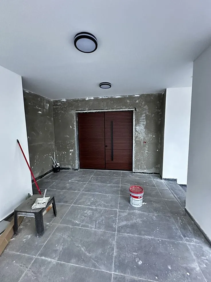 مدخل داخلي أو غرفة قيد التشطيب النهائي، يظهر بها أرضية من البلاط الرمادي، وجدار أمامي يتطلب أعمال اللياسة والطلاء المتقنة التي يوفرها مقاول ترميم الدمام، مع باب خشبي مزدوج باللون البني الداكن مثبت في المنتصف، وإضاءة سقف حديثة مثبتة