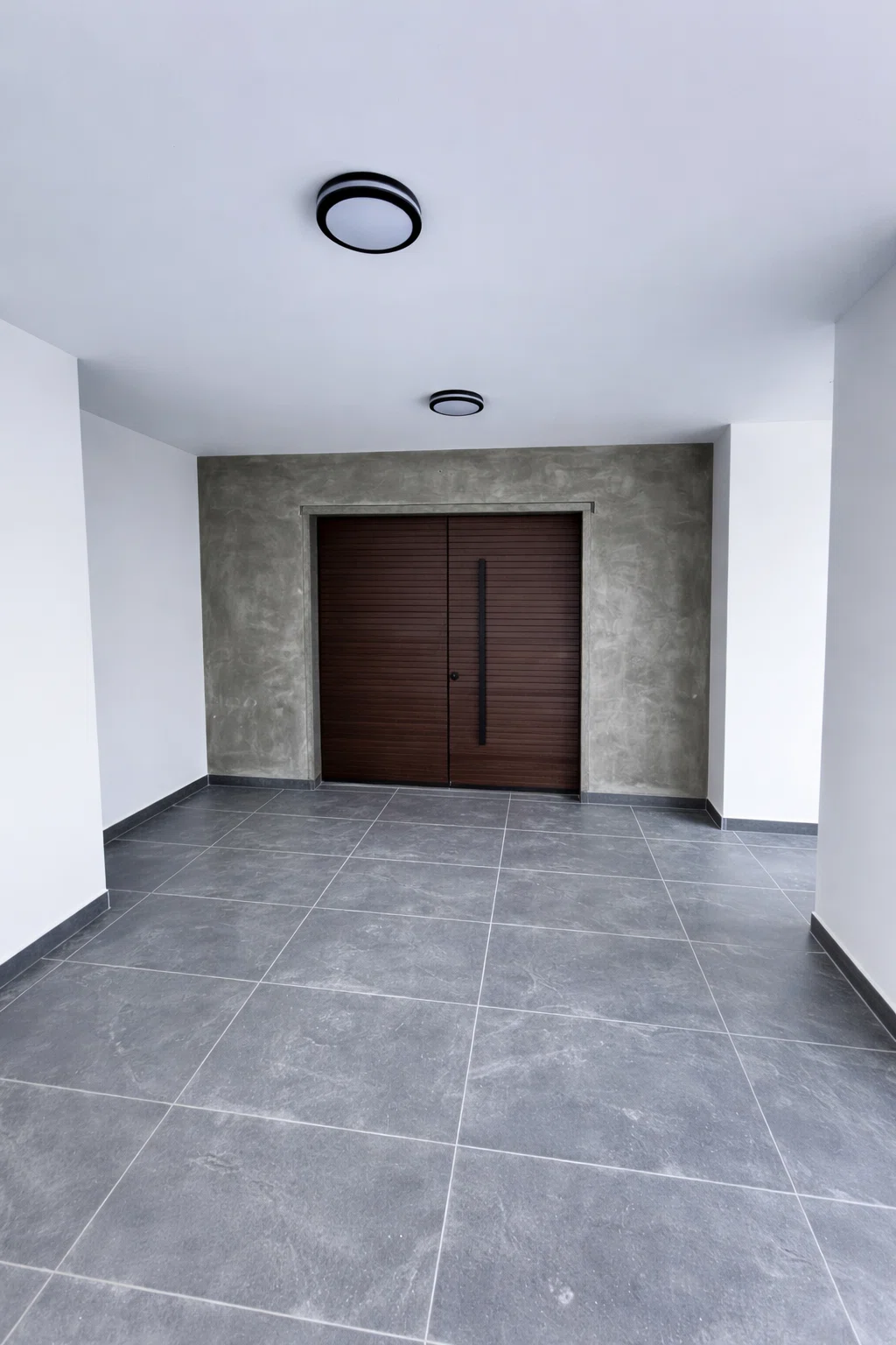 مدخل منزل حديث بعد أعمال الترميم يظهر أرضيات سيراميك رمادية وجدران بتشطيب ديكوري مع باب خشبي بني