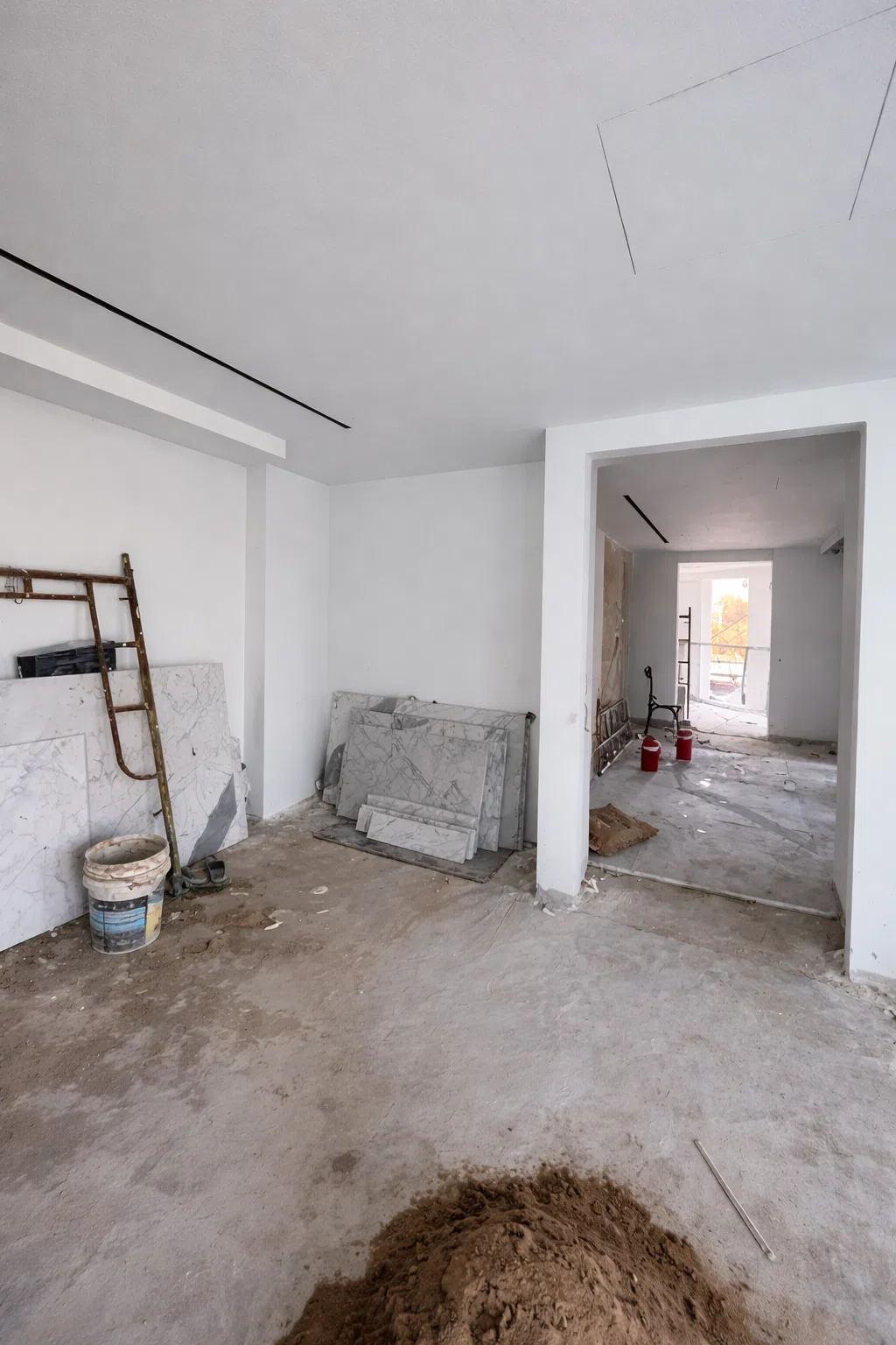 غرفة داخل المنزل أثناء أعمال الترميم تظهر أرضية غير مكتملة وجدران بيضاء مع ألواح رخام وأدوات بناء