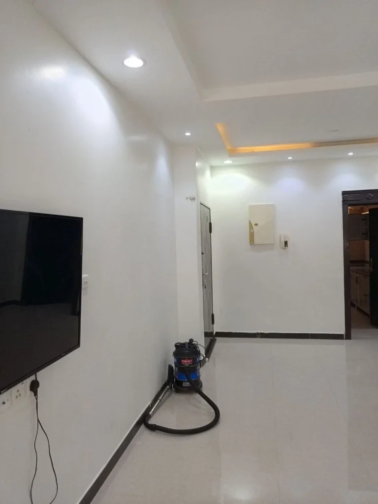 غرفة داخلية بجدران بيضاء وأرضية بلاط رمادي، مع سقف يحتوي على إضاءة على الجدار الأيسر تلفاز مثبت