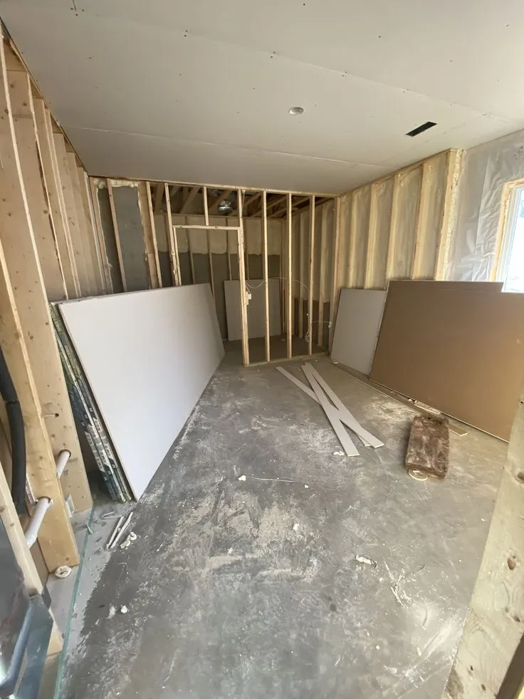 غرفة داخلية في مرحلة إعادة الهيكلة والتقسيم، تظهر فيها الأسقف المركبة وجدران الهياكل الخشبية (Framer) قبل تركيب ألواح الجبس (Drywall)، وهي جزء من أعمال البناء والترميم التي يقوم بها مقاول ترميم الدمام، مع ألواح الجبس جاهزة للتركيب في الموقع