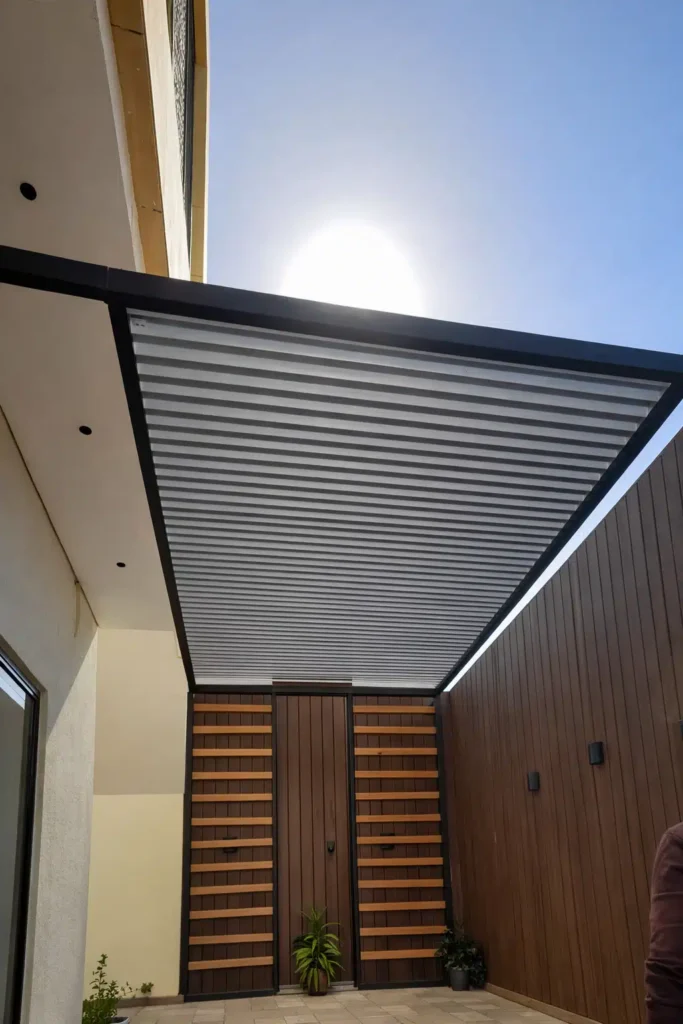 مدخل جانبي بتكسية خشبية عمودية وأبواب بتفاصيل أفقية تغطيه مظلة ألمنيوم شرائحية بإطار أسود مع ظهور الشمس