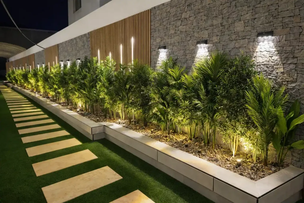 ممر حديقة خارجي ليلي بأرضية عشب صناعي وألواح حجرية متدرجة، يجاوره حوض زراعي طويل مزروع بنباتات خضراء مع إضاءة أرضية
