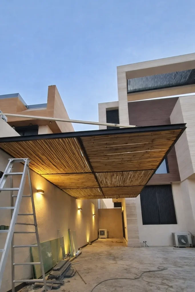 ساحة جانبية بين مبنيين حديثين يغطيها سقف مظلة بإطار معدني أسود وتكسية خشبية طبيعية مع إضاءة جدارية دافئة