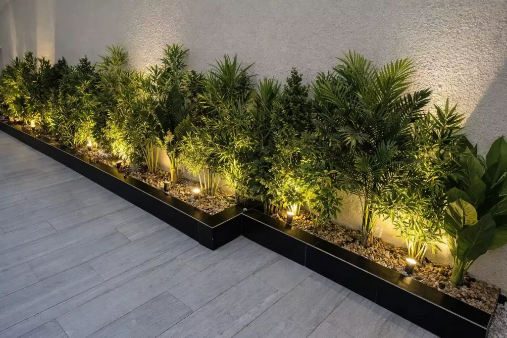 حوض نباتات ملاصق للجدار يضم شجيرات خضراء متنوعة مع سبوتات أرضية بإضاءة تعكس ظلال النباتات على الجدار وأرضية خارجية رمادية