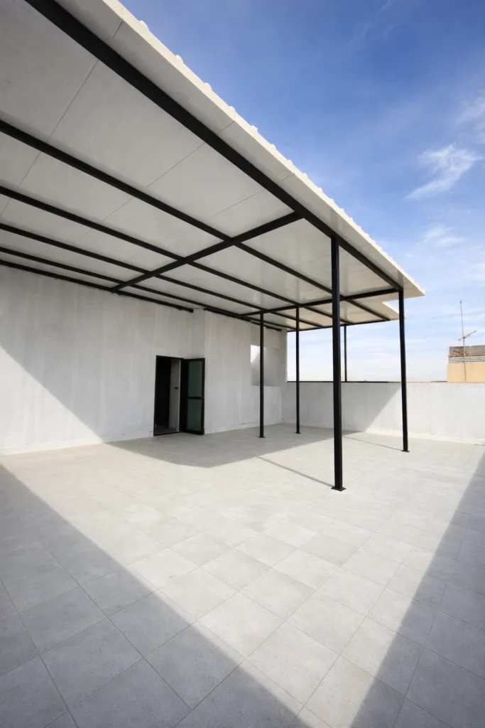 مظلة سطح من الساندويتش بانل بهيكل حديدي أسود فوق مساحة خارجية واسعة بأرضيات بلاط وجدران بيضاء