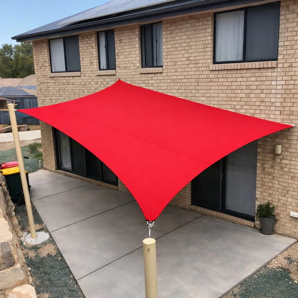 مظلة شد قماشية حمراء مشدودة بأربع زوايا فوق ساحة خلفية أمام منزل بواجهة طوب فاتح مع أعمدة تثبيت معدنية وأرضية خرسانية مكشوفة