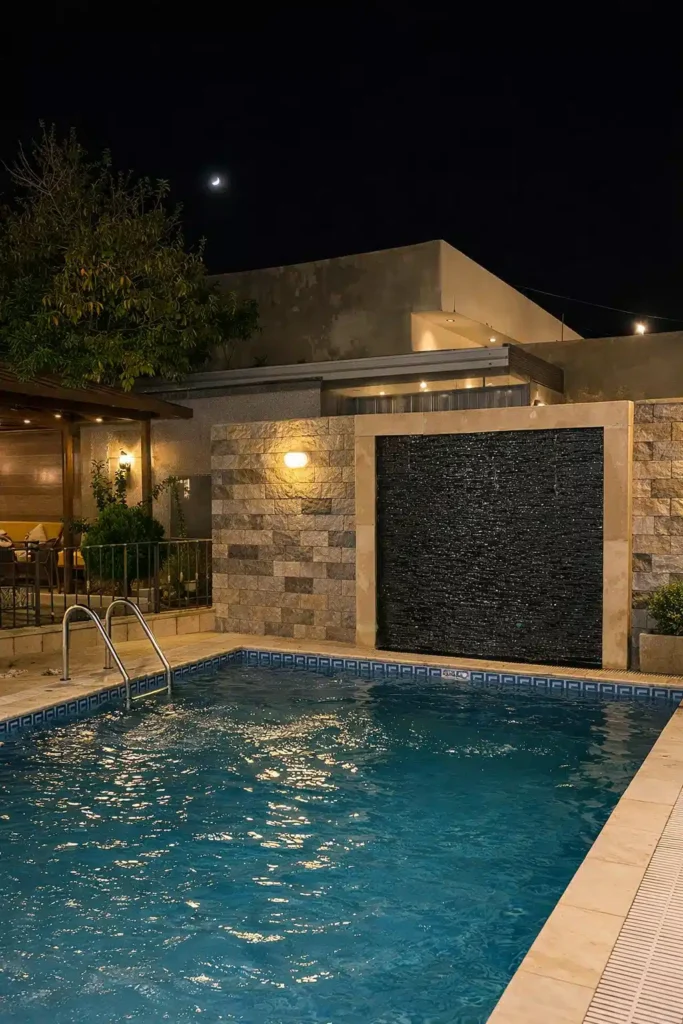 مسبح خارجي منزلي بإضاءة ليلية، يتوسط ساحة خاصة محاطة بجدران حجرية حديثة مع شلال جداري ينساب إلى الماء، وسلّم جانبي للدخول